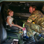 Ein Soldat in militärischer Tarnung steht bei einer kleinen, blonden Schülerin in einem gelben Kleid im Cockpit eines Hubschraubers. Der Soldat lächelt und berührt die Schülerin auf freundliche Weise.