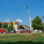 Auf einem Rasen vor einem Gebäude mit Feuerwehrautos steht ein Hubrettich mit Personen, während im Hintergrund Bäume und Straßenlaternen zu sehen sind. Ein Mann geht auf einer Promenade im Vordergrund, und Flaggen an einem Fahnenmast symbolisieren vermutlich eine Veranstaltung.