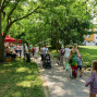 Ein Spaziergangsstreifen führt eine Gruppe von Menschen am Rande eines Marktplatzes vorbei, wo ein Markt mit Ständen stattfindet. Im Hintergrund ist ein grüner Park mit Bäumen und einem Wohngebäude zu sehen.