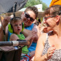 Ein Mann hält ein junges Kind in einem Kinderwagen, während seine Frau ihm zuhört und ihm die Ohren zuhält. Im Hintergrund ist eine Menschenmenge auf einem Fest oder Konzert zu sehen.