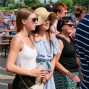 Auf dem Foto sind mehrere Menschen, darunter eine junge Frau mit Strohhut und Sonnenbrille, in einer Gruppe am Ufer eines Sees zu sehen. Im Hintergrund sitzen andere Personen an Tischen und genießen die sommerliche Atmosphäre.