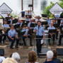 Ein Orchester mit über 30 Musikern in blauen Uniformen spielt auf einer erhöhten Bühne vor Publikum. Ein Dirigent hält die Arme erhoben und spricht während eines Auftritts.