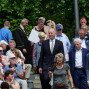Auf einem Treppenaufgang versammelt sich eine große Gruppe von Menschen, darunter Politiker in Anzug und Robe, beim Betrachten einer Ankündigung. Viele Zuschauer sitzen oder stehen umringt von Bäumen, während einige mit Handys fotografieren oder zuhören.