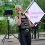 Eine blonde Sängerin singt auf einer Bühne in Schwedt und trägt einen schwarzen Shirt mit der Aufschrift "Paris". Im Hintergrund steht ein Schild mit dem Namen "Stadtsparkasse Schwedt" und eine kleine Bühne.