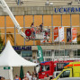 Auf dem Foto ist ein Höhenrettungsgerät auf einem Gebäude montiert, vor dem eine Menschenmenge steht. Im Hintergrund befinden sich ein Rettungsfahrzeug, ein Feuerwehrauto und ein Marktstand mit Werbeplakaten.