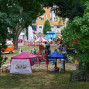 Auf einer Wiese, durch einen dichten Baum voragiert, befindet sich ein Festzelt mit zahlreichen Spielgeräten und Menschen. Im Hintergrund sind einen Geländeraden und ein weiterer Pavillon zu sehen, die das Bild mit einem belebten Festumfeld abrunden.