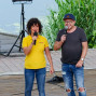 Zwei Personen mit Mikrofonen stehen auf einer Holzterrasse vor einem blauen Lautsprecher und einem grünen Grasstreifen. Eine Frau in gelber Kleidung hält ein Mikrofon, während ein Mann in Jeans und Hemd daneben mit einem Mikrofon spricht.
