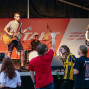 Auf einer Bühne spielen eine Band mit Schlagzeug, Bass und Gitarrist vor einem Publikum. Im Vordergrund stehen mehrere Zuschauer, die die Musik genießt und mit den Händen klatscht.