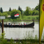 Auf einem ruhigen Wasser fließt ein traditionelles Wikingerschiff mit Touristen an Bord. Im Hintergrund befindet sich ein grüner Wald und eine Fahne, sowie ein hoher Betonpylon.