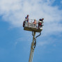 Auf dem Foto sieht man eine Arbeitsplattform in der Höhe, auf der zwei Feuerwehrleute sitzen. Der Himmel ist blau mit weißen Wolken und ein Vogel fliegt im Hintergrund.