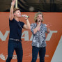 Zwei Musiker singen auf einer Bühne vor einem orangefarbenen Bühnenbild. Sie tragen Kleidung, die für eine Live-Performance geeignet ist, und interagieren mit dem Publikum durch Gesten.