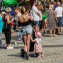 Es ist ein belebter Stadtplatz an einem sonnigen Tag, auf dem eine Gruppe von Menschen, darunter Frauen und Kinder mit Kinderwagen, zusammenkommt. Im Vordergrund steht eine junge Frau, die einen kleinen Mädchenwagen trägt, während um sie herum viele Menschen in lässiger Kleidung stehen und sitzen.